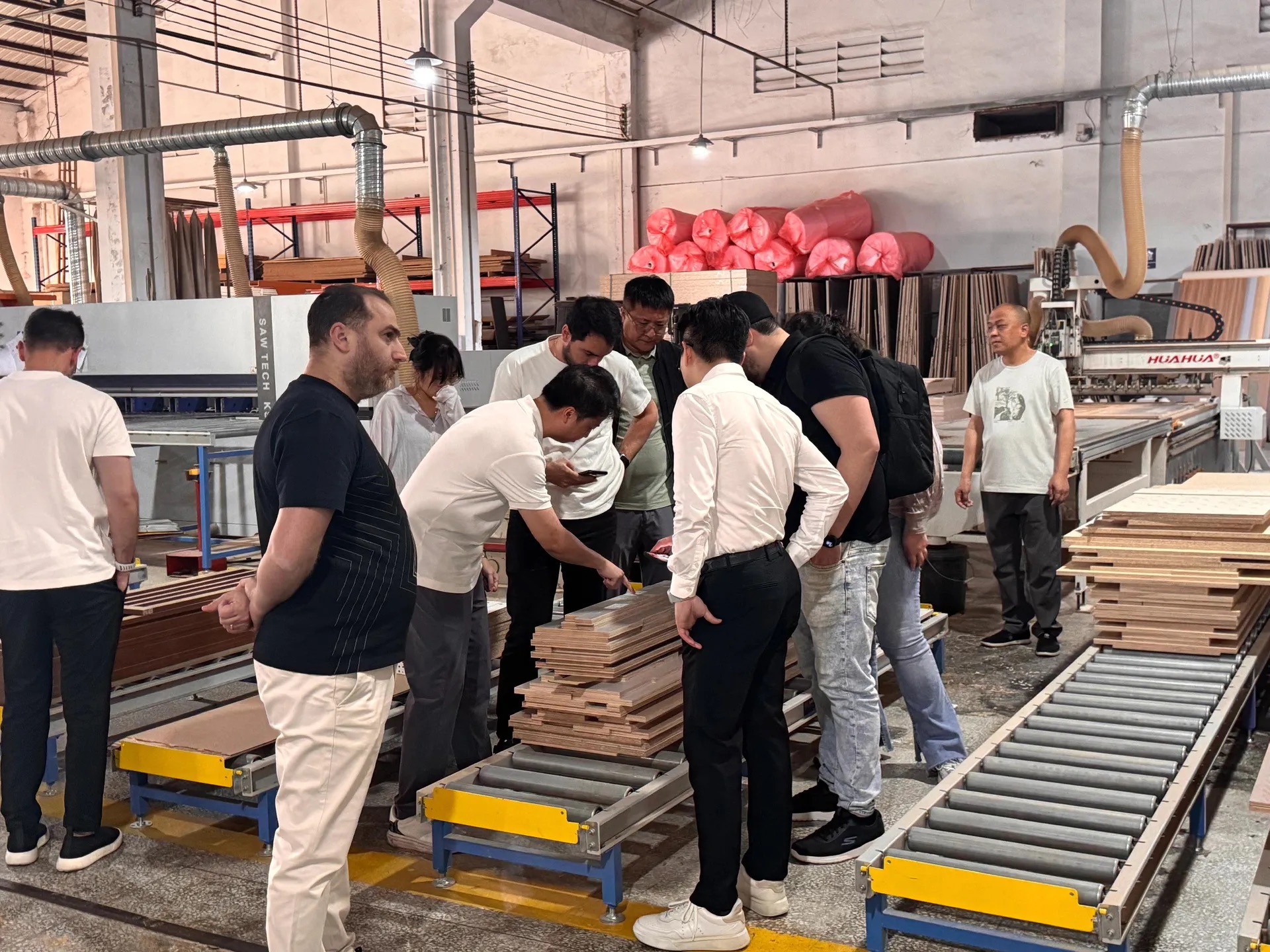 A group reviewing stacked wooden panels beside production machinery in a factory workshop